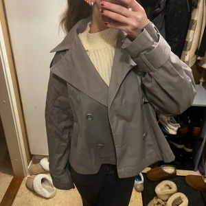 Grå kappa från Gina - Snygg grå kappa från Gina med dubbelknäppning och bred krage. Perfekt för höst och vår. Jackan har en klassisk design med en modern twist och är lätt att matcha med olika outfits. Superfint skick!