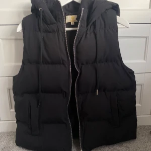 Svart dunväst från Chelsea - Snygg svart dunväst från Chelsea med dragkedja och huva. Använd fåtal gånger, bra skick. Storlek M, liten i storleken och passar S och XS. Säljer för att den inte kommer till användning längre.