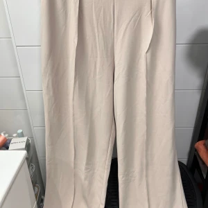 Kostymbyxor från H&M - Snygga beige byxor från H&M i en tight passform. Perfekta för en stilren look med sin vida design. Byxorna har en hög midja och är tillverkade i ett mjukt material som ger en bekväm känsla.