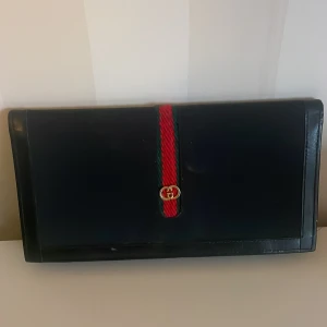 Svart plånbok med röd och grön detalj - Elegant svart plånbok från Gucci med ikonisk röd och grön rand och guldlogotyp. Plånboken har flera fack för kort och en dragkedja för mynt. Perfekt för att hålla ordning på dina värdesaker med stil.