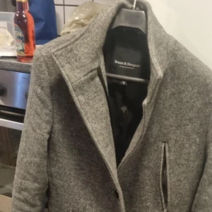 Grå kappa från Bruun & Stengade - Snygg grå kappa från Bruun & Stengade med klassisk design. Perfekt för kyligare dagar med stilren look och bekväm passform. Kappan har en elegant krage och diskreta fickor framtill.