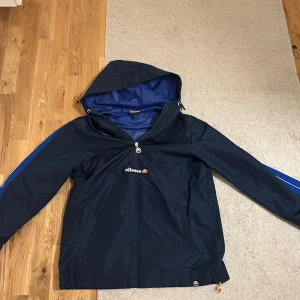 Blå vindjacka från Ellesse - Säljer en blå vindjacka från Ellesse med huva och dragkedja. Jackan har en sportig look med blå ränder längs ärmarna och Ellesse-logga på bröstet. Perfekt för blåsiga dagar.