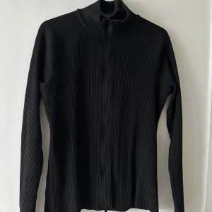 Svart ribbad tröja med dragkedja - Snygg svart ribbad tröja med dragkedja framtill. Perfekt för lager-på-lager och har en stilren design med långa ärmar. 