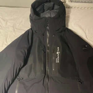 Svart Sail Racing Gore-Tex jacka - Säljer en svart Sail Racing jacka i Gore-Tex material. Jackan har en hög krage och flera praktiska dragkedjor framtill. Perfekt för kalla och blåsiga dagar med sin robusta design och funktionella detaljer. Den har ett litet hål i ryggen som går lätt att sy och är ändå nästan osynbar.