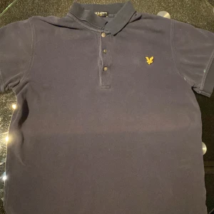Mörkblå pikétröja från Lyle & Scott - Säljer en klassisk mörkblå pikétröja från Lyle & Scott med gul broderad logga på bröstet. Tröjan har korta ärmar och tre knappar framtill. Perfekt för en stilren look. Använd i 1 månad, ej kvitto
