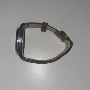 Klocka med brun läderrem från GANT - Stilren klocka från GANT med en brun läderrem och en silverfärgad boett. Urtavlan är vit med tydliga markeringar och en subdial för extra funktionalitet. Perfekt för en klassisk look. Bilden där klockan är spend är på den första pluppen/ spänningen. Det är budgivning och minsta priset är 500 kr