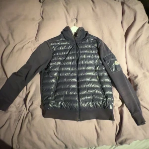Svart jacka från Moncler - Snygg svart jacka från Moncler med quiltad framsida och dragkedja. Jackan har långa ärmar och en praktisk ficka med knapp på ärmen. Perfekt för kyligare dagar. Är lite solblekt som syns på bilderna men några skador