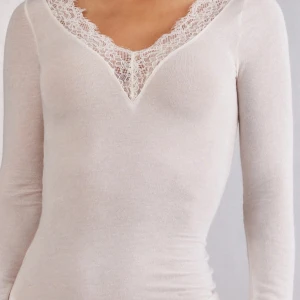 Vit långärmad topp med spetsdetaljer från intimissimi - Elegant vit långärmad topp med v-ringning och spetsdetaljer vid hals och ärmslut. Perfekt för en stilren look.