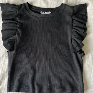 Svart ribbad topp med volangärmar från Zara - Snygg svart ribbad topp från Zara med volangdetaljer på ärmarna. Perfekt för en trendig look. Toppen har en rund halsringning och är i en bekväm passform.
