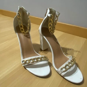 Vita sandaler med guldkedjor - Stilrena vita sandaler med klack och dekorativa guldkedjor. Sandalerna har en dragkedja baktill för enkel på- och avtagning. Perfekta för att ge en elegant touch till din outfit.