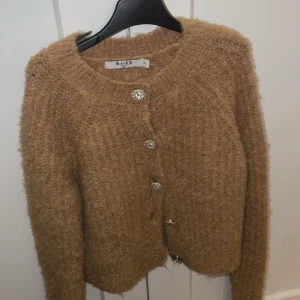 Beige kofta från NA-KD - Mysig beige kofta från NA-KD med glittriga knappar. Perfekt för kyliga dagar med sin fluffiga textur och långa ärmar. En stilren och bekväm tröja som passar till många tillfällen.