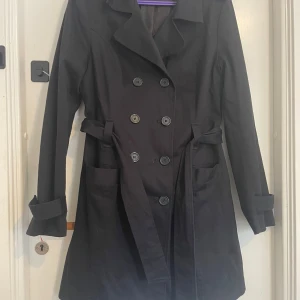 Svart trenchcoat från Guess Jeans - Säljer en snygg svart trenchcoat från Guess Jeans. Den har en klassisk dubbelknäppt design med ett bälte i midjan för en stilren look. Jackan är långärmad och perfekt för höst och vår. Den är i bra skick och redo för nya äventyr!
