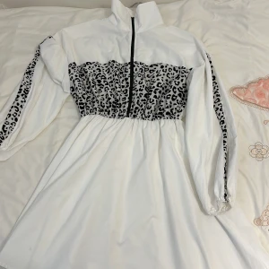 Vit klänning med leopardmönster - Säljer en supercool vit klänning med leopardmönster på bröstet och längs ärmarna. Den har en dragkedja framtill och hög krage, vilket ger en sportig look. Klänningen är i ett lätt och bekvämt material, perfekt för en avslappnad dag eller en festlig kväll. Den har en normal passform och ärmlängden är lång.