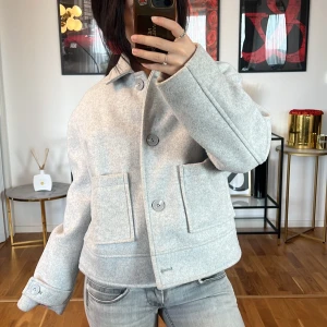 Ljusgrå ulljacka - Säljer en superfin ljusgrå jacka i ullmaterial. Den har en klassisk krage och stora knappar framtill. Jackan är lite croppad och har stora fickor som ger en cool look. Perfekt för höst och vår när du vill vara både stilren och varm. Passar perfekt till jeans för en avslappnad stil.