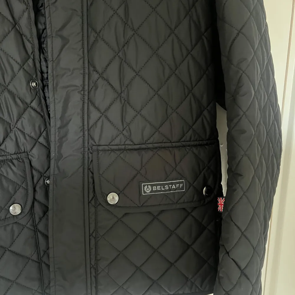 Säljer en snygg svart quiltad jacka från Belstaff. Jackan har en stilren design med långa ärmar och praktiska fickor framtill. Den är perfekt för höst och vår, och har både dragkedja och knappar för stängning. Jackan är i nyskick och redo för nya äventyr!. Jackor.