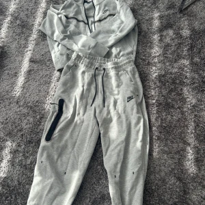 Grå mjukisbyxor från Nike - Säljer ett par grå mjukisbyxor och hoodie från Nike (tjejmodellen). De är superbekväma och perfekta för chill dagar eller träning. Byxorna har en elastisk midja med snörning och en cool svart dragkedja på sidan. Säljer för att de är för små för mig. 