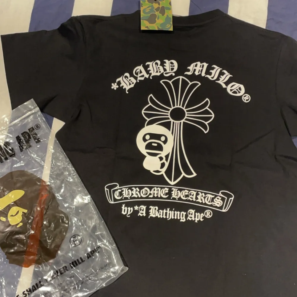 Säljer en svart t-shirt från det coola samarbetet mellan Chrome Hearts och A Bathing Ape. T-shirten har ett stort tryck med 'Chrome Hearts' och 'Baby Milo' i vitt på både fram- och baksidan. Den är kortärmad och perfekt för en avslappnad stil. Taggen är i camo-mönster vilket ger en extra touch.. T-shirts.