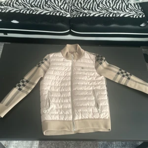 Beige quiltad jacka från Burberry - Säljer en snygg beige quiltad jacka från Burberry. Den har ett klassiskt rutigt mönster på ärmarna och baksidan, och en dragkedja framtill. Jackan är perfekt för höst och vår, med en stilren look som passar både till vardags och fest. Den är i nyskick och redo för en ny ägare!