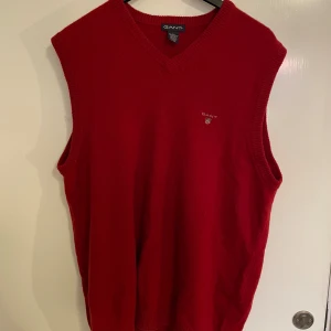 Röd stickad väst från GANT - Säljer en snygg röd stickad väst från GANT. Den har en klassisk v-ringad design och är perfekt för lager-på-lager under höst och vinter. Västen är i storlek XL och är i bra skick. Perfekt för en stilren och bekväm look!