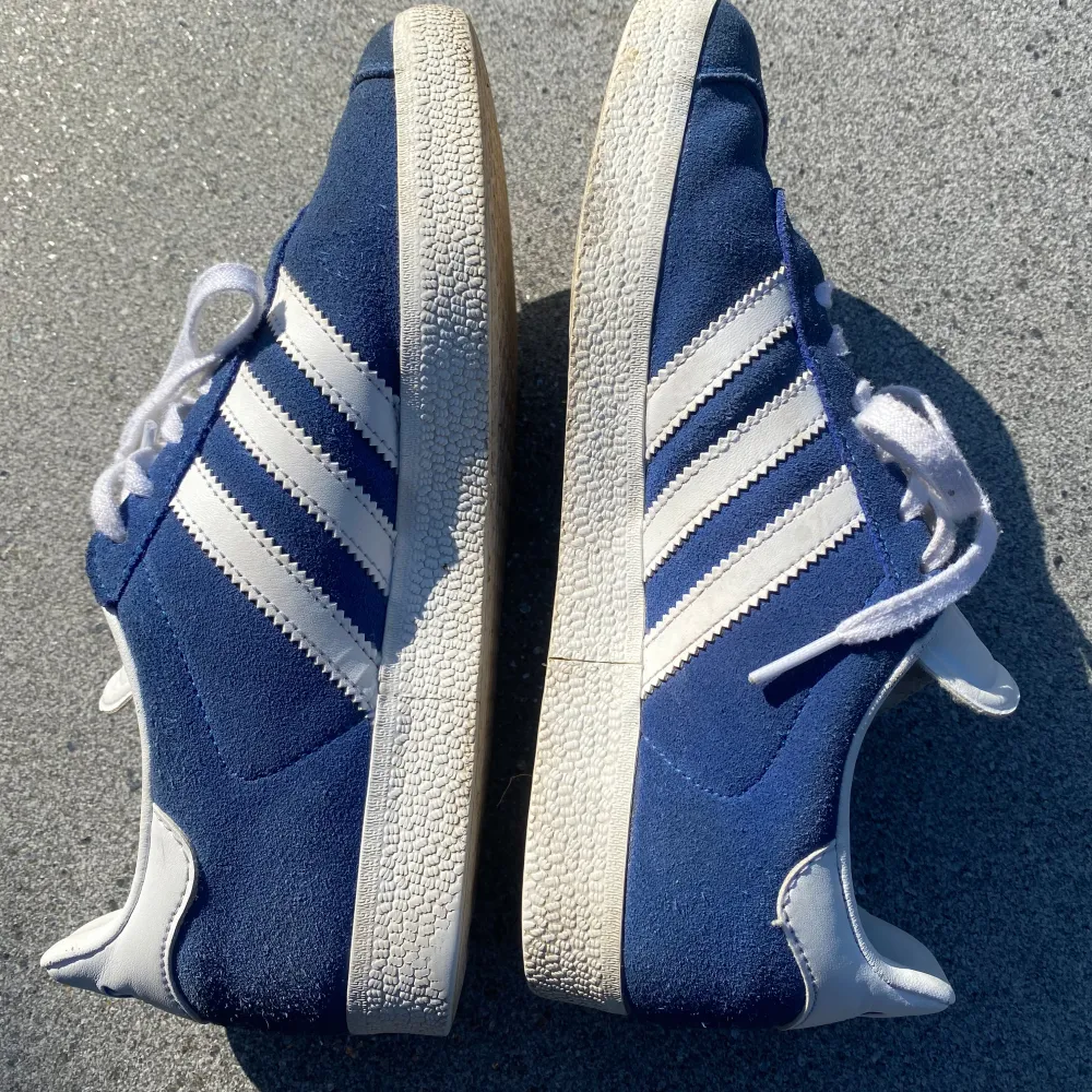 Säljer ett par klassiska blå Adidas Gazelle sneakers i mocka med vita detaljer. Skorna har den ikoniska tre-randiga designen och vit sula. De är i mycket bra skick med lite slitage på sulan. Perfekta för både vardag och fest! . Skor.