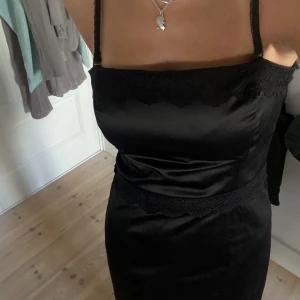 Svart axelbandslös klänning - Säljer en svart klänning med spetsdetaljer. Den har justerbara axelband som också är avtagninsbara, vilket gör den perfekt för både fest och mer formella tillfällen. Klänningen är i ett glansigt material som ger en lyxig känsla, assnygg med en blazer