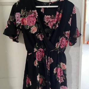 Svart blommig klänning med knytband - Säljer en superfin svart playsuit med ett vackert blommönster i rosa och grönt. Playsuiten har en omlott-design med knytband i midjan och korta ärmar. Perfekt för sommarens fester eller en mysig vårdag!