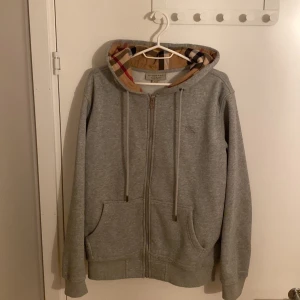 Grå huvtröja från Burberry - Säljer en snygg grå huvtröja från Burberry med klassiskt rutigt mönster i huvan. Tröjan har dragkedja framtill och är i nyskick. Perfekt för höst och vår! Tröjan är i storlek L men passar för dom som har M också.