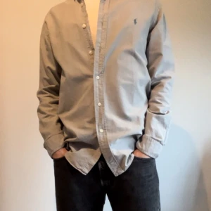 Grå skjorta från Ralph Lauren M - Säljer en stilren grå skjorta från Ralph Lauren i slim fit. Skjortan har långa ärmar och knappar framtill. Perfekt för både vardag och fest! 🕺     Storlek M (på bild: 176cm)        Nypris 1499kr och skjortan är fri från defekter och är i mycket bra skick!
