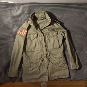 Säljer en snygg olivgrön jacka från Ralph Lauren med coola detaljer som en amerikansk flagga på ärmen och flera praktiska fickor. Jackan har en klassisk krage och är perfekt för höst och vår. Den är i bra skick och passar perfekt för en casual look.