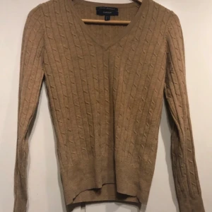 Beige stickad tröja från Gant - Säljer en superfin beige stickad tröja från Gant i stretchig lammull. Den har en snygg v-ringning och ett klassiskt kabelstickat mönster. Perfekt för höst och vinter, och den är i bra skick, finns några noppor och ett litet hål. Se bilder. Passar både till vardags och lite finare tillfällen. 🧶✨