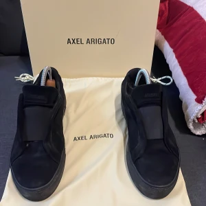 Svarta sneakers från Axel Arigato - Säljer ett par stilrena svarta sneakers från Axel Arigato i mycket bra skick. Skorna är i skinn och har en minimalistisk design utan snörning. Perfekta för både vardag och fest. Kommer med originalpåse och kartong.