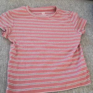 Rosa randig t-shirt från H&M - Säljer en söt rosa randig t-shirt från H&M Basic. Den har korta ärmar och är gjord i ett mjukt bomullsmaterial. Perfekt för vardagsbruk och passar bra till både jeans och shorts. T-shirten är i mycket bra skick och redo för en ny ägare!