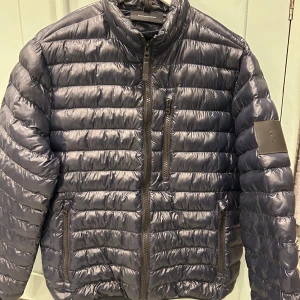 Svart pufferjacka från Peak Performance - Säljer en snygg glansig mörkblå pufferjacka från Peak Performance. Jackan har en dragkedja framtill och flera praktiska fickor och är i riktigt bra skick endast använd några gånger. Perfekt för kyliga dagar och håller dig varm och bekväm. Passar både till vardags och utomhusaktiviteter.
