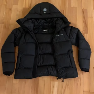Svart dunjacka från Sail Racing - Säljer en svart dunjacka från Sail Racing i storlek medium. Jackan är i mycket bra skick och har en fyllning av 700 fill power dun från Allied Feather + Down. Perfekt för kalla vinterdagar med sin varma och bekväma design. Den har en dragkedja framtill och en justerbar huva. Texten är lite borta med man ser vad det står fortfarande.