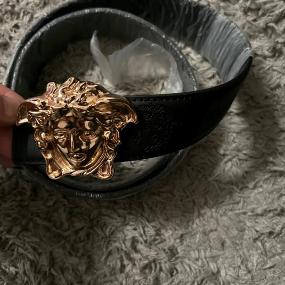 Säljer ett snyggt svart skärp från Versace med ett stort, detaljerat guldspänne i form av Medusa. Skärpet är i skinn och har en lyxig känsla, perfekt för att lyfta vilken outfit som helst. Passar både till fest och vardag. 🖤✨. Övrigt.