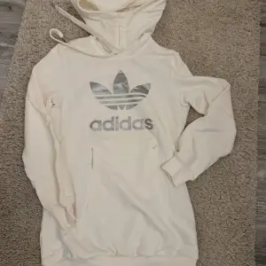 Säljer en snygg vit hoodie från Adidas i storlek 36. Den har en stor silverfärgad logga på framsidan och en praktisk magficka. Perfekt för en avslappnad stil och passar bra till både vardag och träning. Huvan har dragsko för justering. Skön och stilren design!