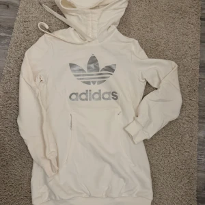 Vit hoodie från Adidas - Säljer en snygg vit hoodie från Adidas i storlek 36. Den har en stor silverfärgad logga på framsidan och en praktisk magficka. Perfekt för en avslappnad stil och passar bra till både vardag och träning. Huvan har dragsko för justering. Skön och stilren design!