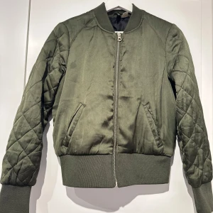 Olivgrön bomberjacka - Säljer en snygg olivgrön bomberjacka i mycket bra skick, endas en liten fläck på framsidan. Jackan har quiltade ärmar och en dragkedja framtill. Perfekt för höst, med ribbade muddar vid ärmslut och nederkant för extra komfort. Passar både till vardags och lite mer uppklädda tillfällen.