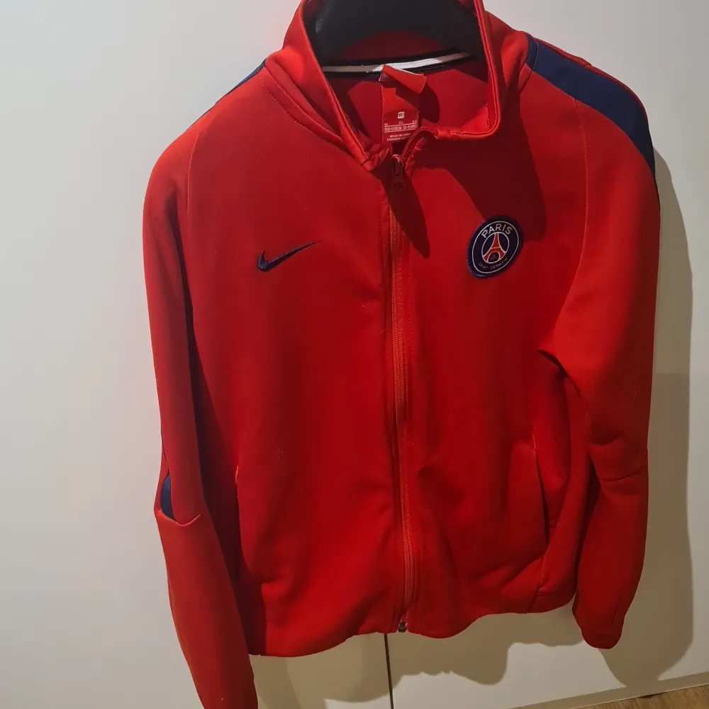 Säljer en snygg röd Nike jacka med PSG-logga på bröstet. Jackan har en dragkedja framtill och långa ärmar. Perfekt för fotbollsfans som vill visa sitt stöd för Paris Saint-Germain. Den är i bra skick och passar perfekt för höst och vår.. Jackor.