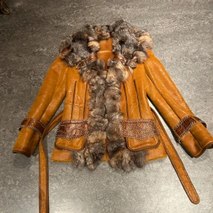 Pälsjacka  - Säljer en  brun pälsjacka med äkta päls från Italien. Jackan har ett bälte i midjan och snygga spännen vid ärmsluten. Perfekt för höst och vinter när du vill hålla dig varm och stilren. Den har en unik design med päls runt kragen och längs framsidan. Passar perfekt för en chic och trendig look!