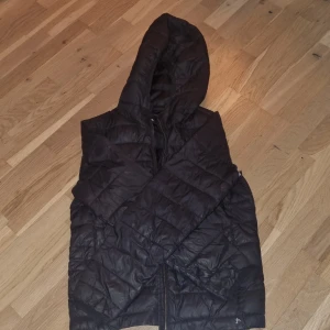 Svart dunväst från Pull&Bear - Säljer en svart jacka från Pull&Bear i storlek S. Jackan har en dragkedja och är perfekt för hösten. Den är quiltad och har en huva för extra värme. Notera att det finns en reva på sidan som behöver lagas. Perfekt för dig som gillar en cool och avslappnad stil!