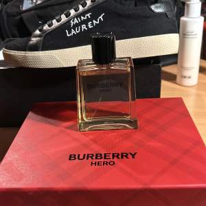 Säljer en elegant Burberry Hero parfym i en stilren, fyrkantig glasflaska med svart kork. Flaskan står på en röd kartong med Burberry-loggan. Perfekt för den som vill ha en sofistikerad doft med en touch av lyx. Passar både till vardags och speciella tillfällen.