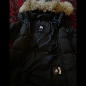 Svart dunjacka canada goose - Säljer en svart dunjacka från Canada Goose, den är i storlek xs och är i bra skick. Säljer då jag vill köpa en längre modell. Dragkedjan flärpen finns tyvärr inte kvar men det går att stänga jackan. Avtagbar päls.  På 4 bilden kan man se hur det är ett litet hål vid jackan men inget man märker av. Andvänd men bra skick. 
