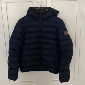 Mörkblå dunjacka från Colmar - Säljer en snygg mörkblå dunjacka från Colmar i storlek 12Y (154 cm). Jackan har en praktisk huva och dragkedja framtill. Perfekt för kalla dagar med sin varma och bekväma passform. Den är i mycket bra skick och redo för nya äventyr!