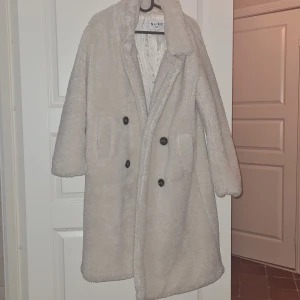 Beige teddyjacka från NA-KD - Mysig beige teddyjacka från NA-KD i mycket bra skick. Perfekt för höst och vinter med sin varma och fluffiga design. Jackan har långa ärmar och stängs med knappar framtill. Den är både stilren och bekväm, idealisk för kyliga dagar.