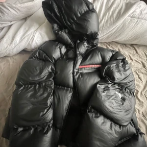 Svart pufferjacka från Prada - Säljer en svart pufferjacka från Prada i storlek M. Jackan är gjord av 100% återvunnen polyester och är vattentät. Den har en stor huva och dragkedja framtill. Perfekt för kalla vinterdagar! Notera att det finns en liten reva på ena ärmen.
