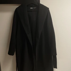 Svart kappa från Zara - Säljer en elegant svart kappa från Zara i väldigt bra skick. Den har en stilren design med knappar och ett bälte i midjan för en snygg passform.  Den har också en luva. Längden är till under knäna (är 164cm lång). Perfekt för höst och vinter! 🖤