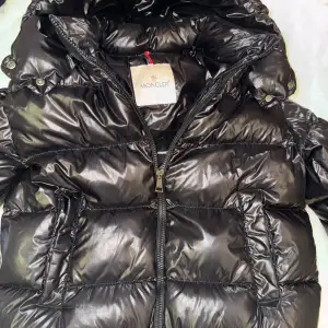 Säljer en snygg svart pufferjacka från Moncler i mycket bra skick. Jackan har en glansig finish och dragkedja framtill. Perfekt för kalla vinterdagar med sin varma vaddering. Den har en unik design med en ljusare detalj på . Passar perfekt för vintern❄️