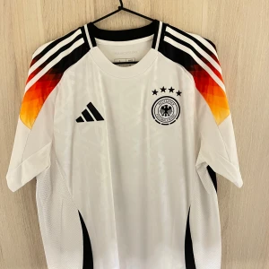 Tysklands hemma tröja 23/24 - Säljer en vit fotbollströja från Adidas med Tysklands landslagsemblem och tre stjärnor på bröstet. Tröjan har korta ärmar och coola färgdetaljer i svart, rött och gult på axlarna. Namnet 'Sané' och nummer 19 pryder ryggen. Perfekt för fotbollsfans! Bara hängt på väggen ej använd. Köpt från unisport butiken i Stockholm i somras 