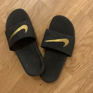 Säljer ett par svarta Nike sandaler i bra skick. De har en snygg guldlogga på remmen och en bekväm sula med bra grepp. Perfekta för sommardagar eller att glida runt hemma. Storlek är inte angiven, men de ser ut att passa de flesta.